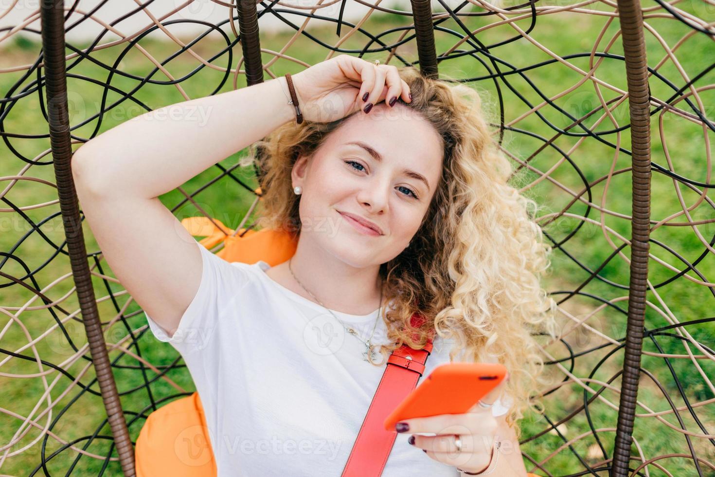 Beautiful delighted female blogger with curly light hair, uses smart phone for posting new posts on her web site, rests in hanging chair, uses wireless internet connection. People, recreation concept photo
