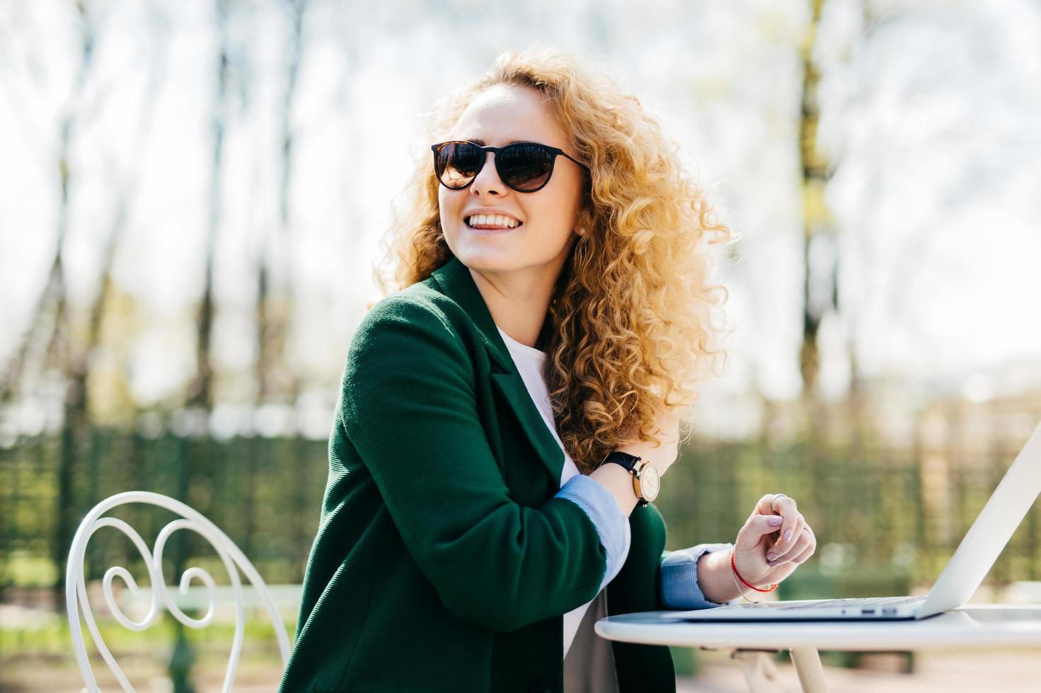 Happy stylish woman with curly light hair wearing sunglasses working with laptop outside in park typing necessary documents turning back noticing someone at the street. Cheerful woman having rest photo
