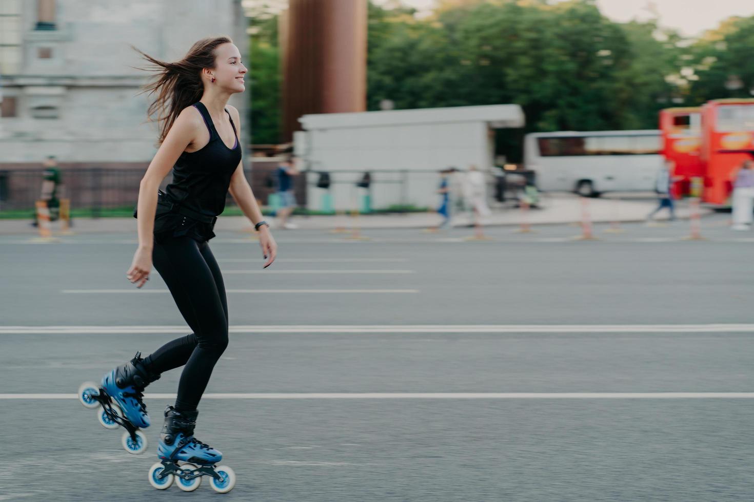 enérgica delgada joven mujer sonriente patines en la carretera de la ciudad disfruta pasar el tiempo libre se mueve activamente rápidamente tiene el pelo oscuro flotando en el viento lleva un estilo de vida saludable. fines de semana activos. foto al aire libre