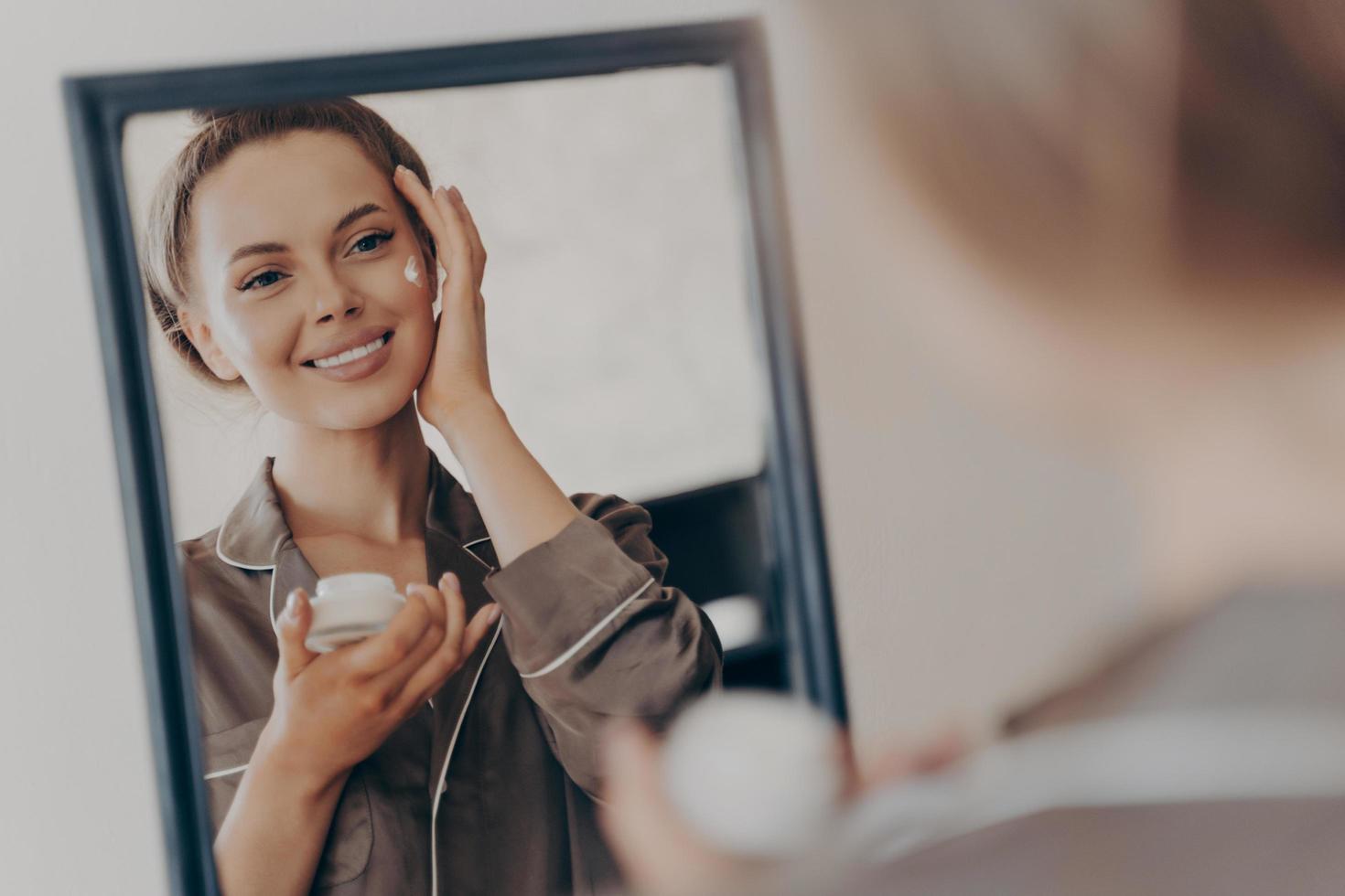 mujer joven positiva y feliz sonriendo mientras aplica crema facial foto