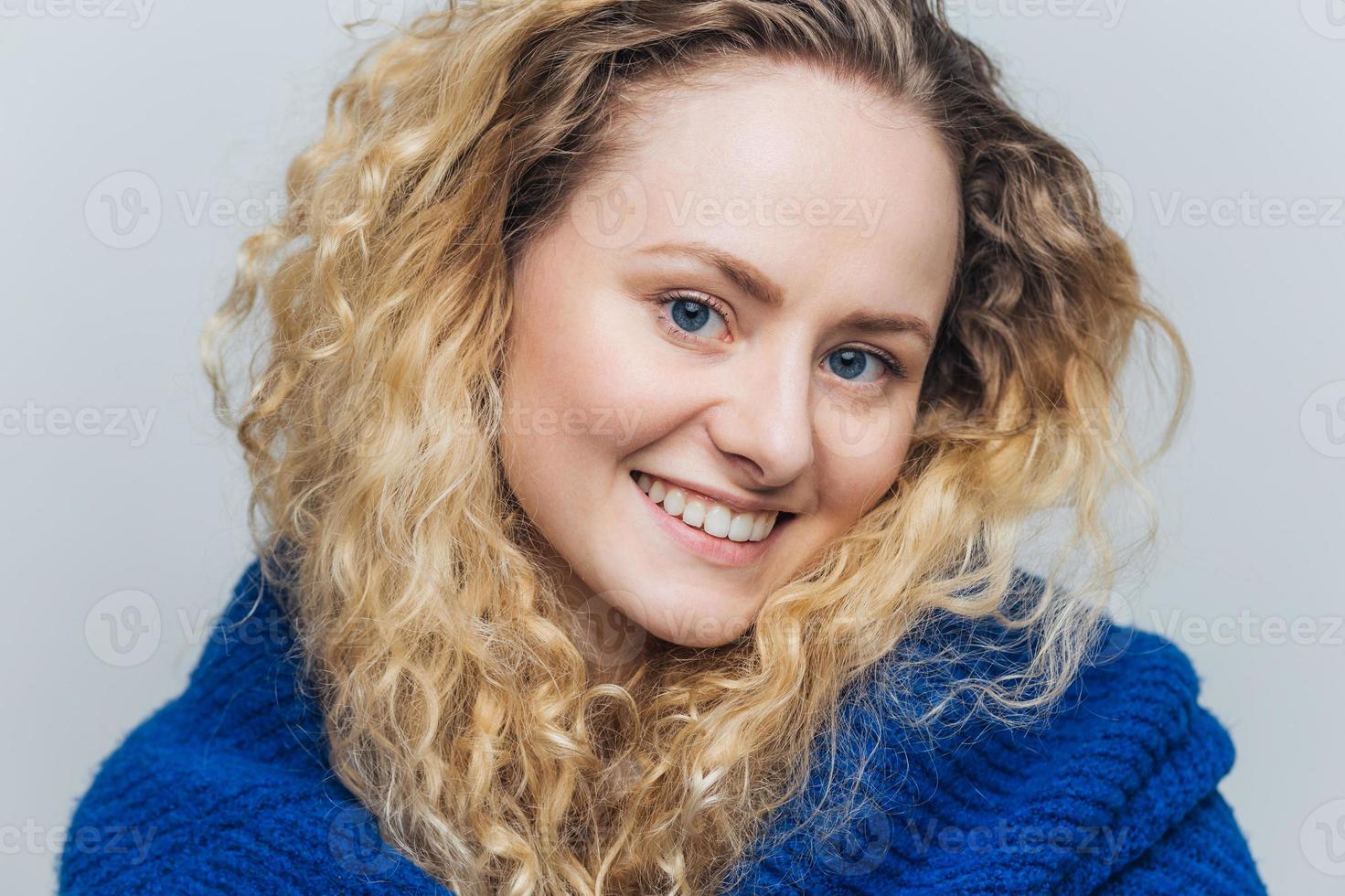 Close up shot of attractive young woman with light crisp hair, blue eyes and shining smile, looks positively at camera, being in good mood after walk outdoor, isolated over light blue background photo