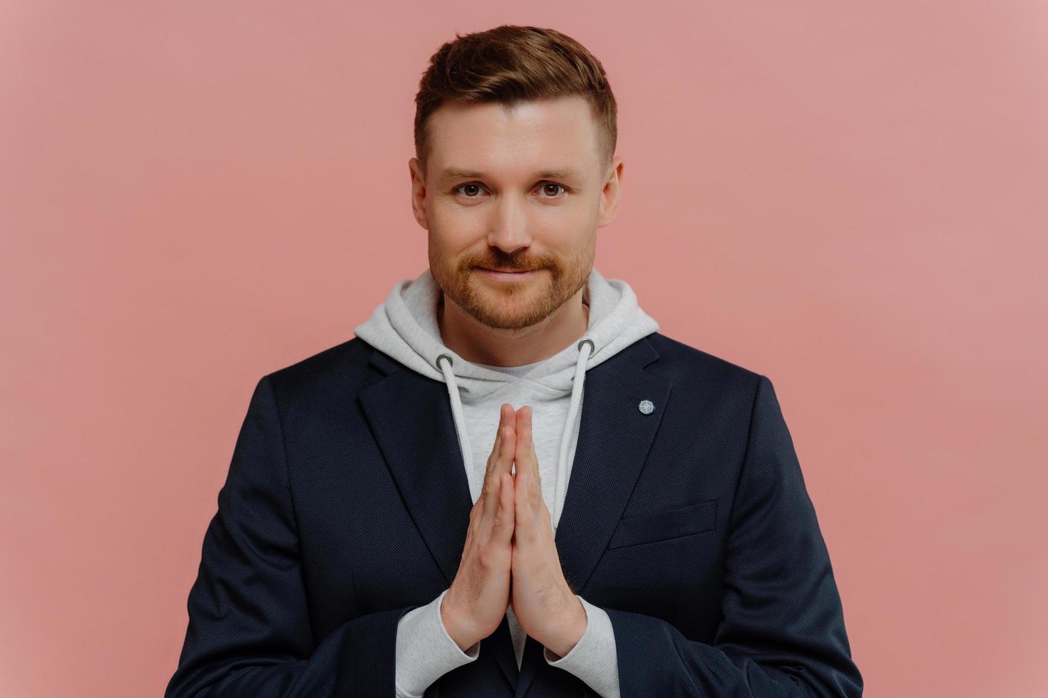 Serious bearded adult man keeps palms pressed together prays for something looks directly at camera believes in something good happen poses against pink background. Handsome guy makes request photo