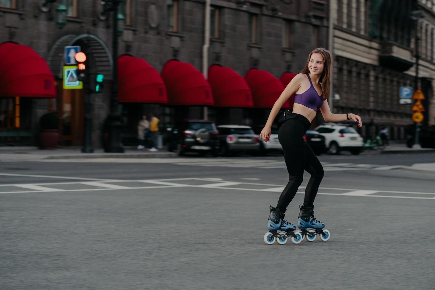 Outdoor shot of active slim woman dressed in cropped top and leggings rollerblades through city enjoys excellent physical workout burns calories gets energy relieves stress learns new skills photo