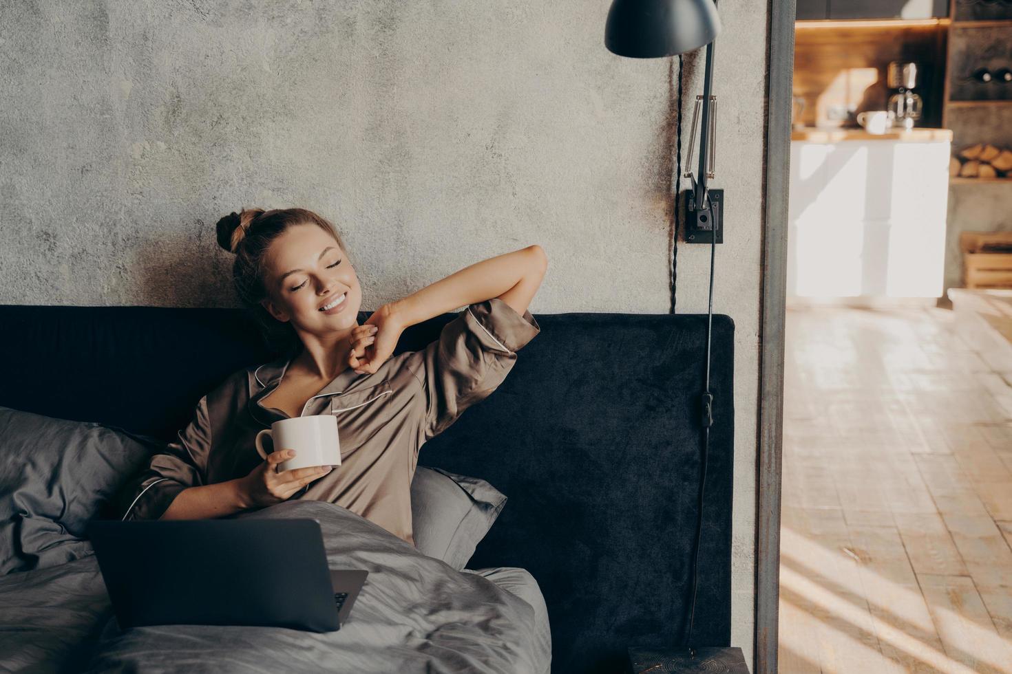 Happy beautiful female with closed eyes drinking coffee in bed before remote work at home photo