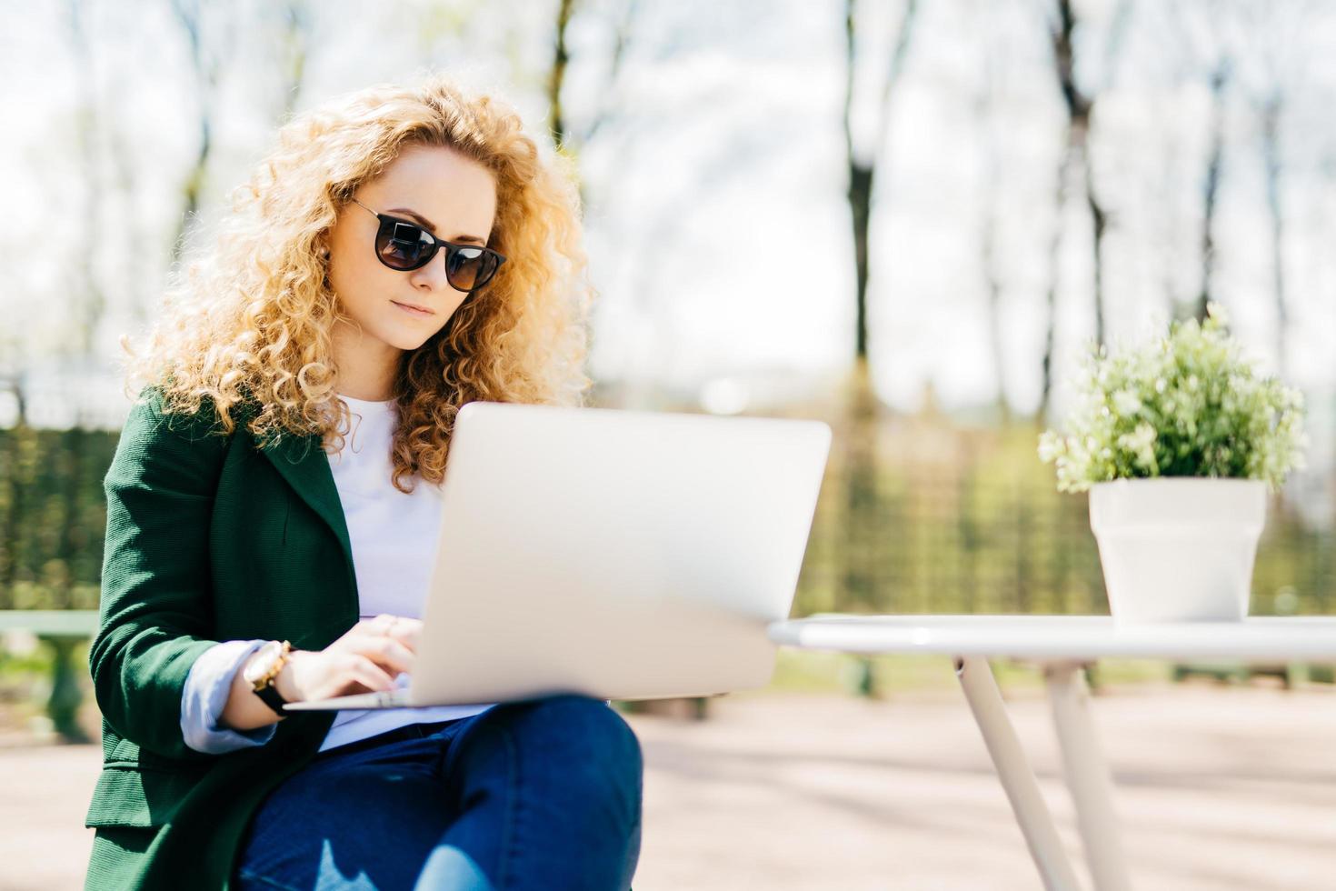 Outdoor portrait of attractive young girl with fluffy blonde hair wearing sunglasses, jeans and green jacket holding laptop computer on her knees reading interesting book online and messaging photo