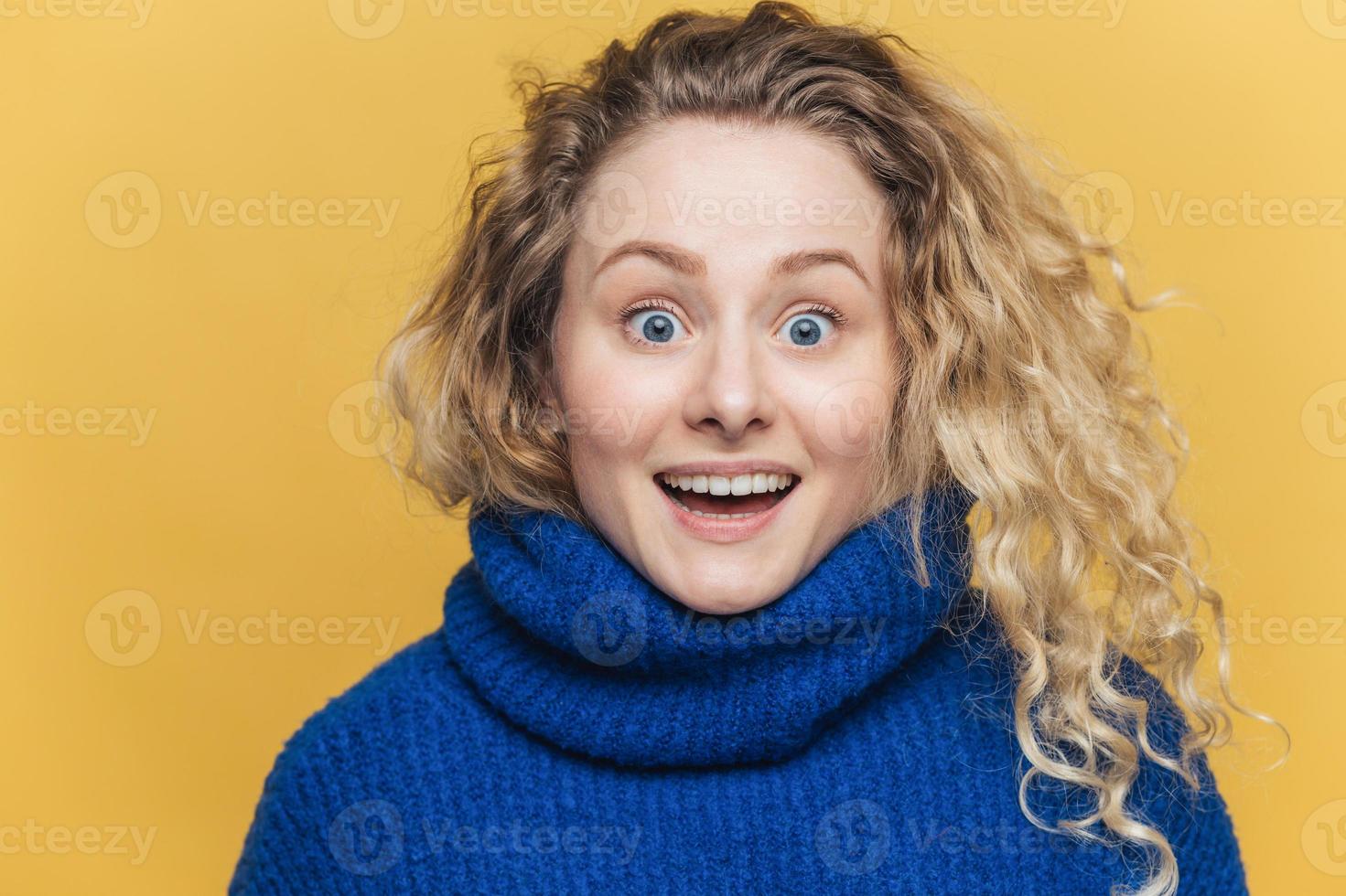Stunned beautiful blue eyed young woman with light crisp hair, doesnt expect to recieve shocking news, wears knitted sweater, stares at camera with bugged eyes, isolated over yellow background photo