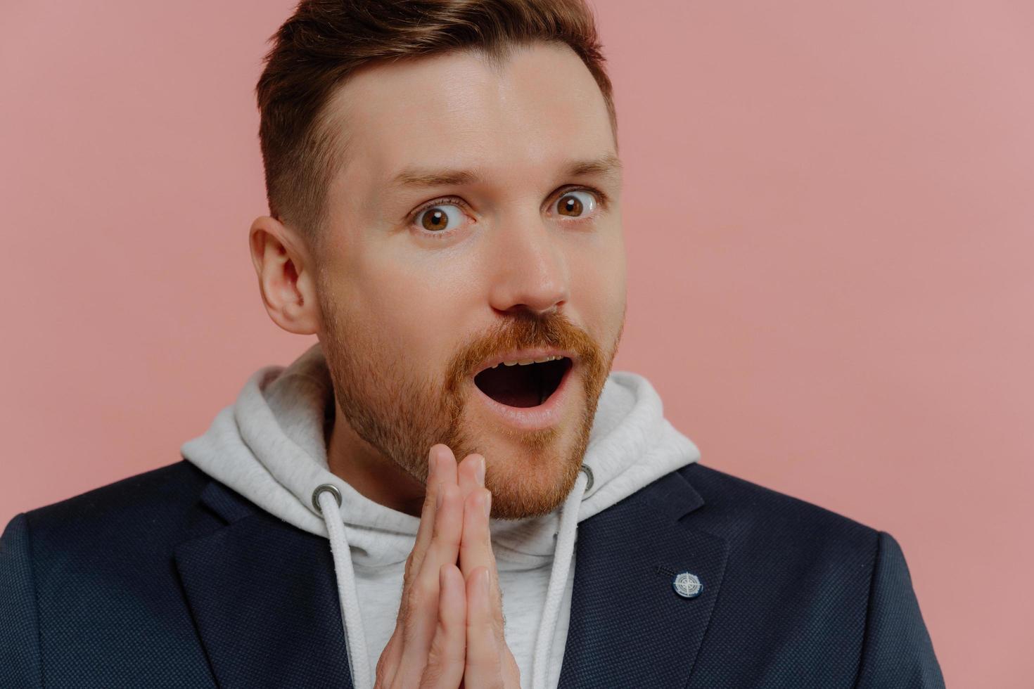 Close up portrait of amazed bearded young European man keeps mouth opened palms pressed together cannot believe in something wears sweatshirt with hood black jacket isolated over pink background photo