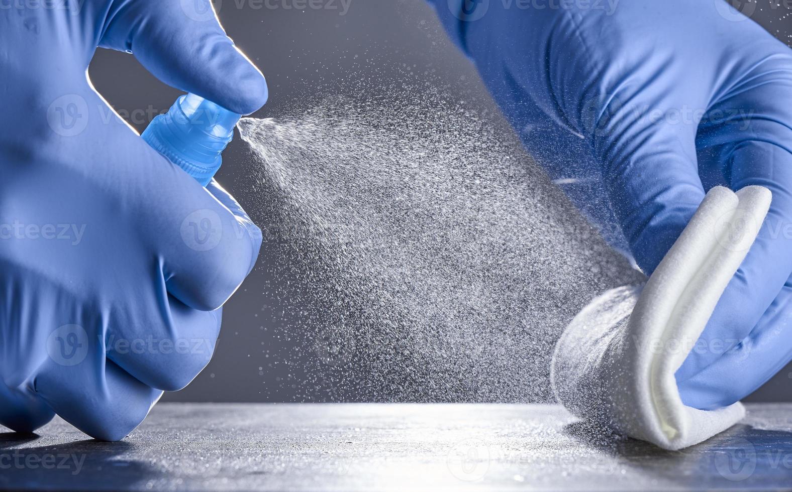 Hands in rubber gloves treat the table with a disinfectant spray on a grey background. Disinfection of premises photo