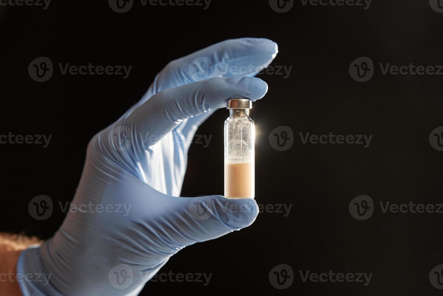 A doctor holds a vial full of blue liquid with his latex gloves on. Isolated on black. photo