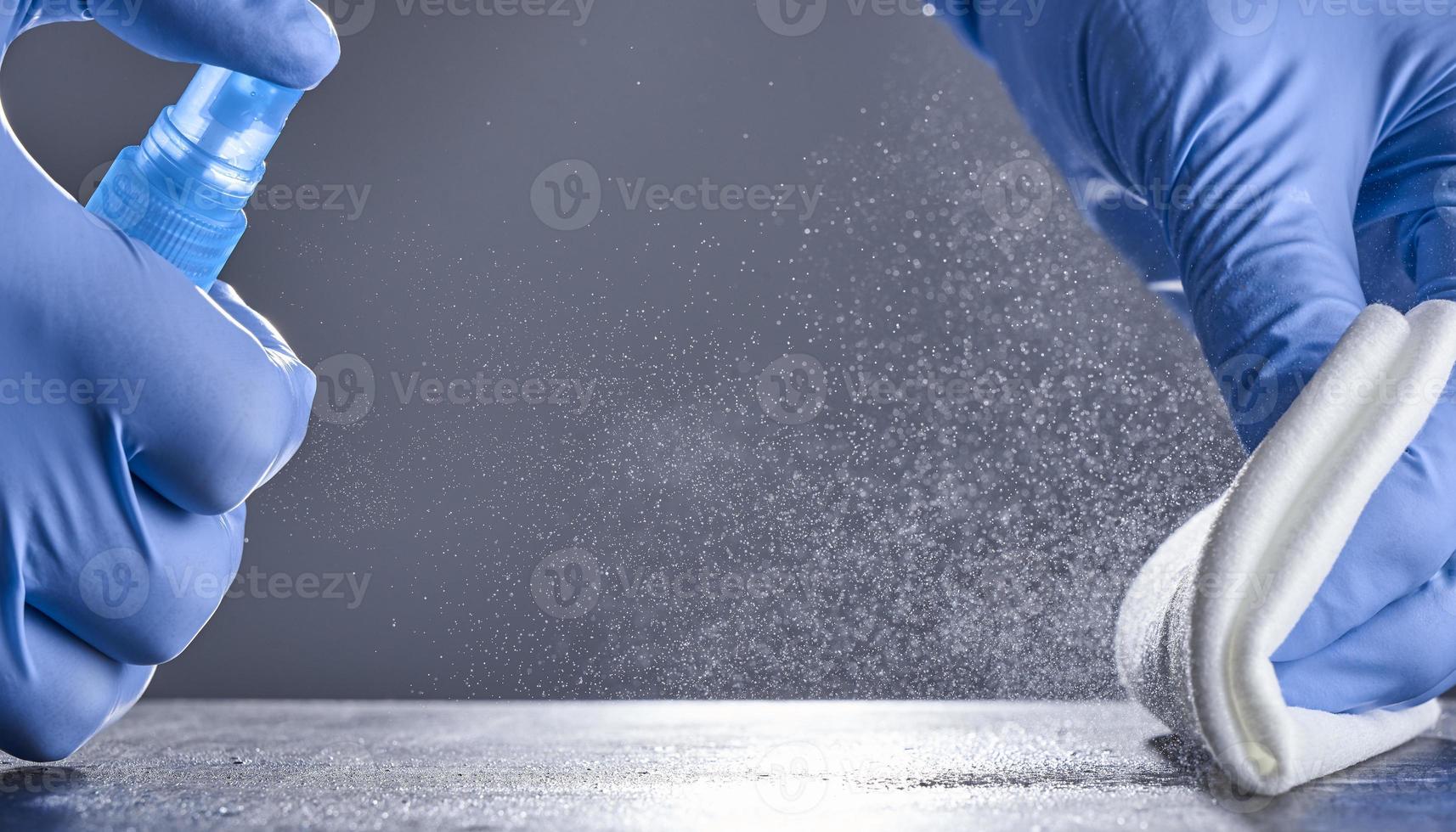 las manos con guantes de goma limpian la mesa con un spray desinfectante sobre un fondo gris. desinfección de locales foto