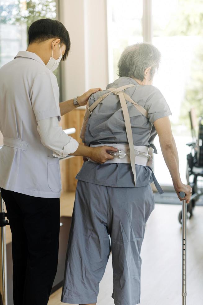 joven fisioterapeuta asiático que trabaja con un anciano usando un andador en el pasillo de un hogar de ancianos foto