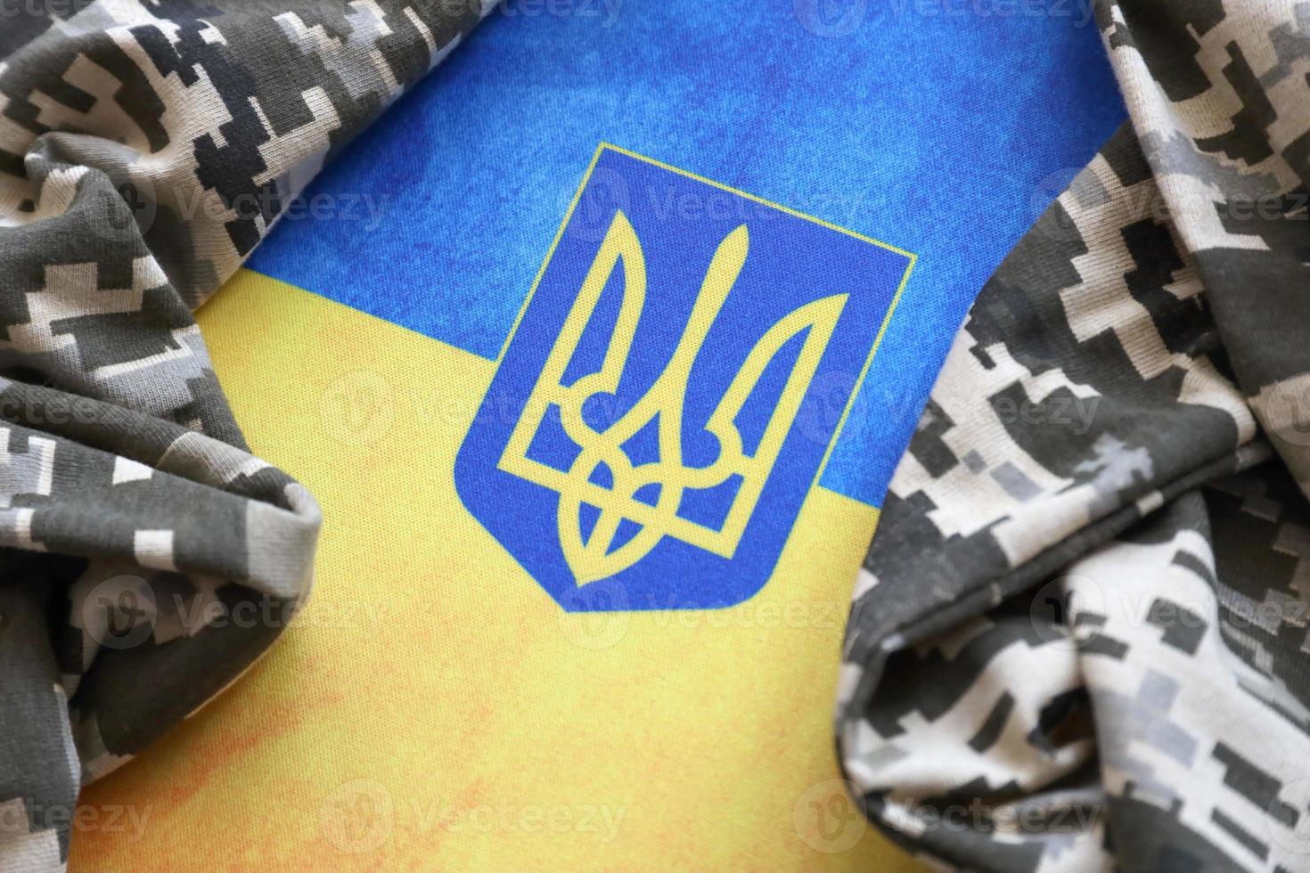 Ukrainian flag and coat of arms with fabric with texture of pixeled camouflage. Cloth with camo pattern in grey, brown and green pixel shapes with Ukrainian trident sign photo