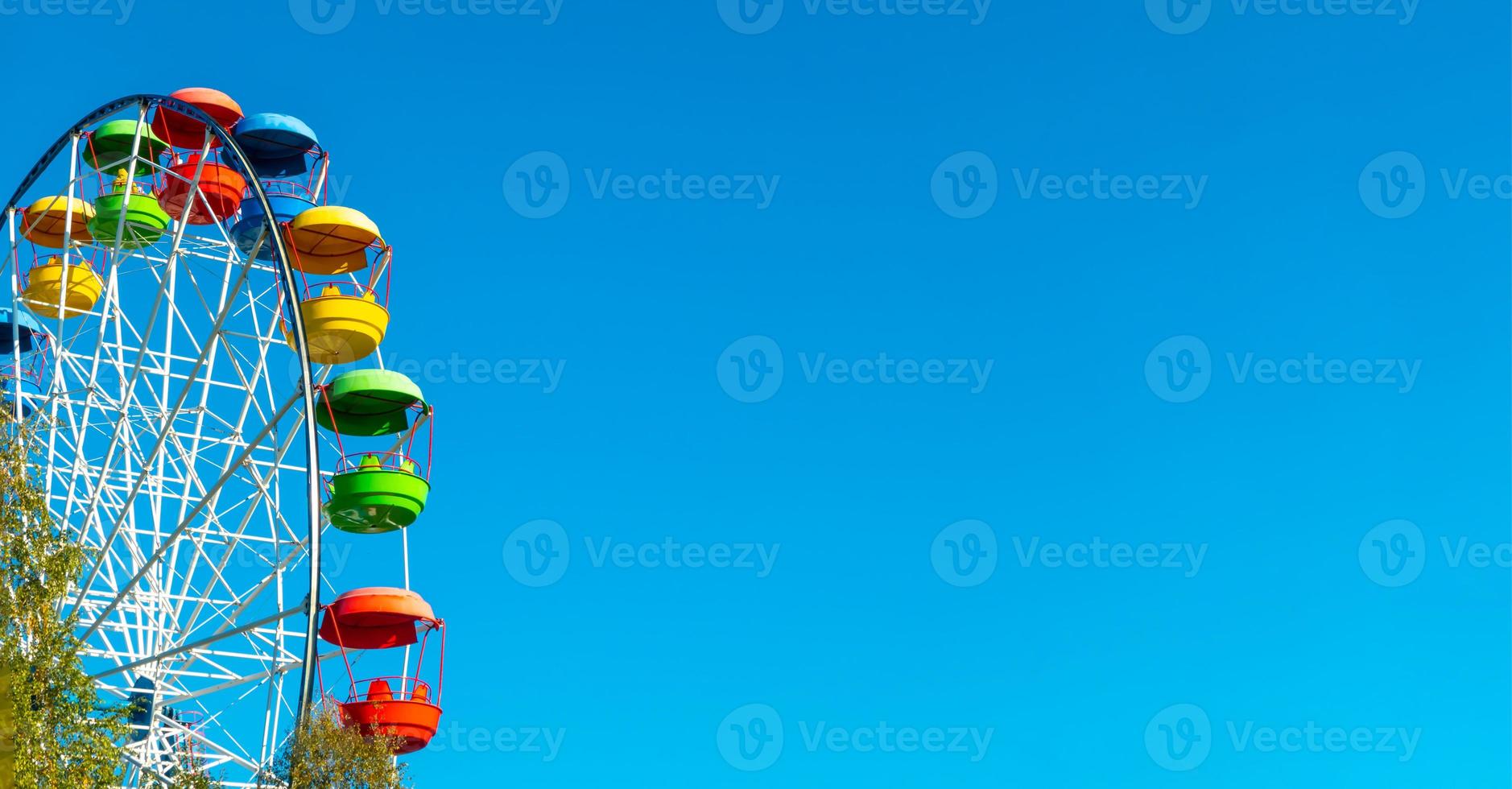 landscape of an amusement park with the top of a Ferris wheel showing above the tree tops against a blue sky. photo