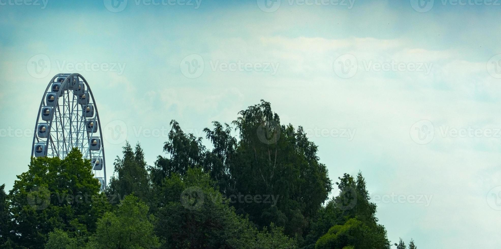 landscape of an amusement park with the top of a Ferris wheel showing above the tree tops against a blue sky. photo