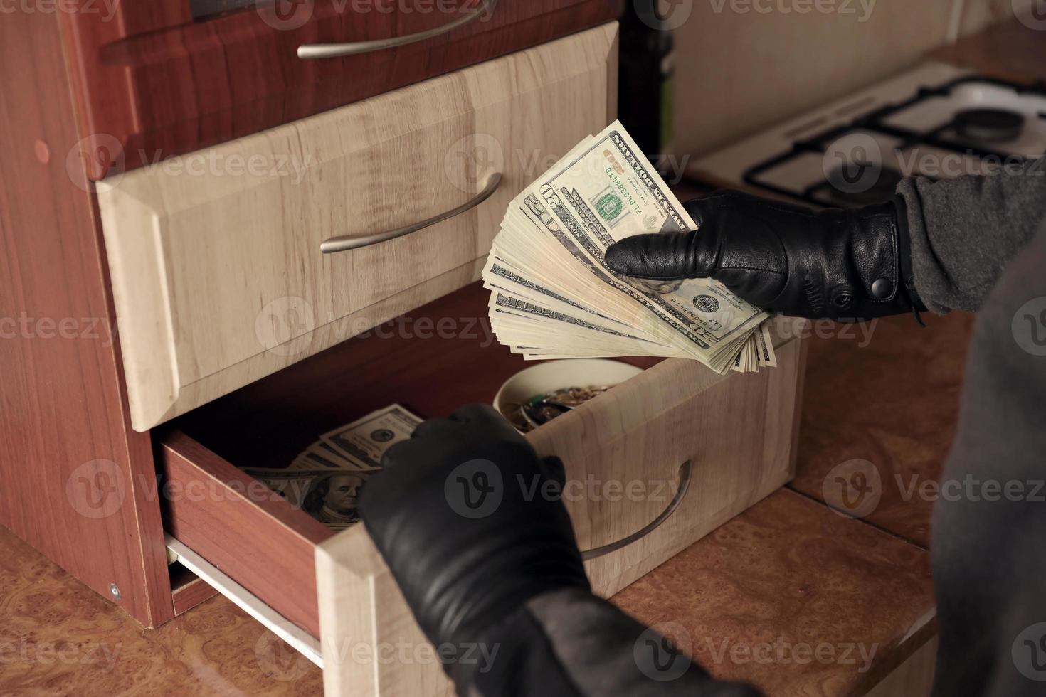 ladrón con traje negro y guantes se ve en un estante abierto en la cocina. el ladrón saca los billetes de dólar estadounidense de un estante foto