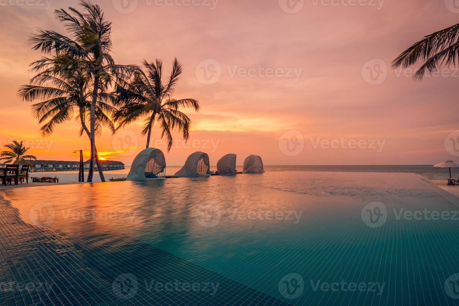 Luxury sunset over infinity pool in a summer beachfront hotel resort at tropical landscape. Tranquil beach holiday vacation background mood. Amazing island sunset beach view, palms swimming pool photo