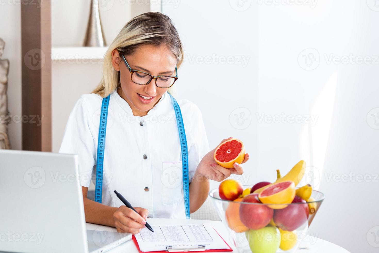 Portrait of young smiling female nutritionist in the consultation room. Nutritionist desk with healthy fruit, juice and measuring tape. Dietitian working on diet plan. photo