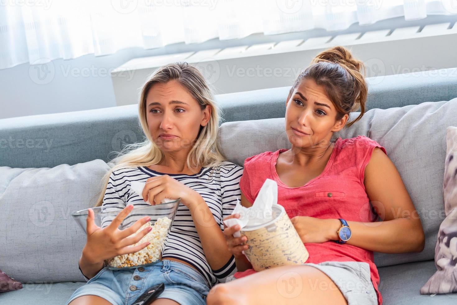 two best friends watch sad movie at home in bed crying touching. girls hold popcorn and tv remote control looking screen romantic film on television. ladies in pajama wiping tear using tissue photo