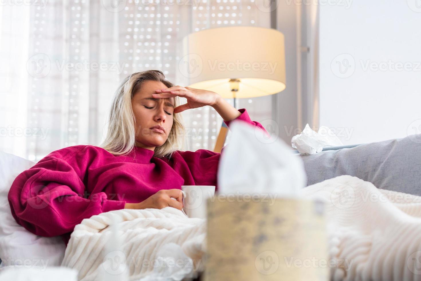 Sick woman with headache sitting under the blanket. Sick woman with seasonal infections, flu, allergy lying in bed. Sick woman covered with a blanket lying in bed with high fever, Coronavirus symptoms photo