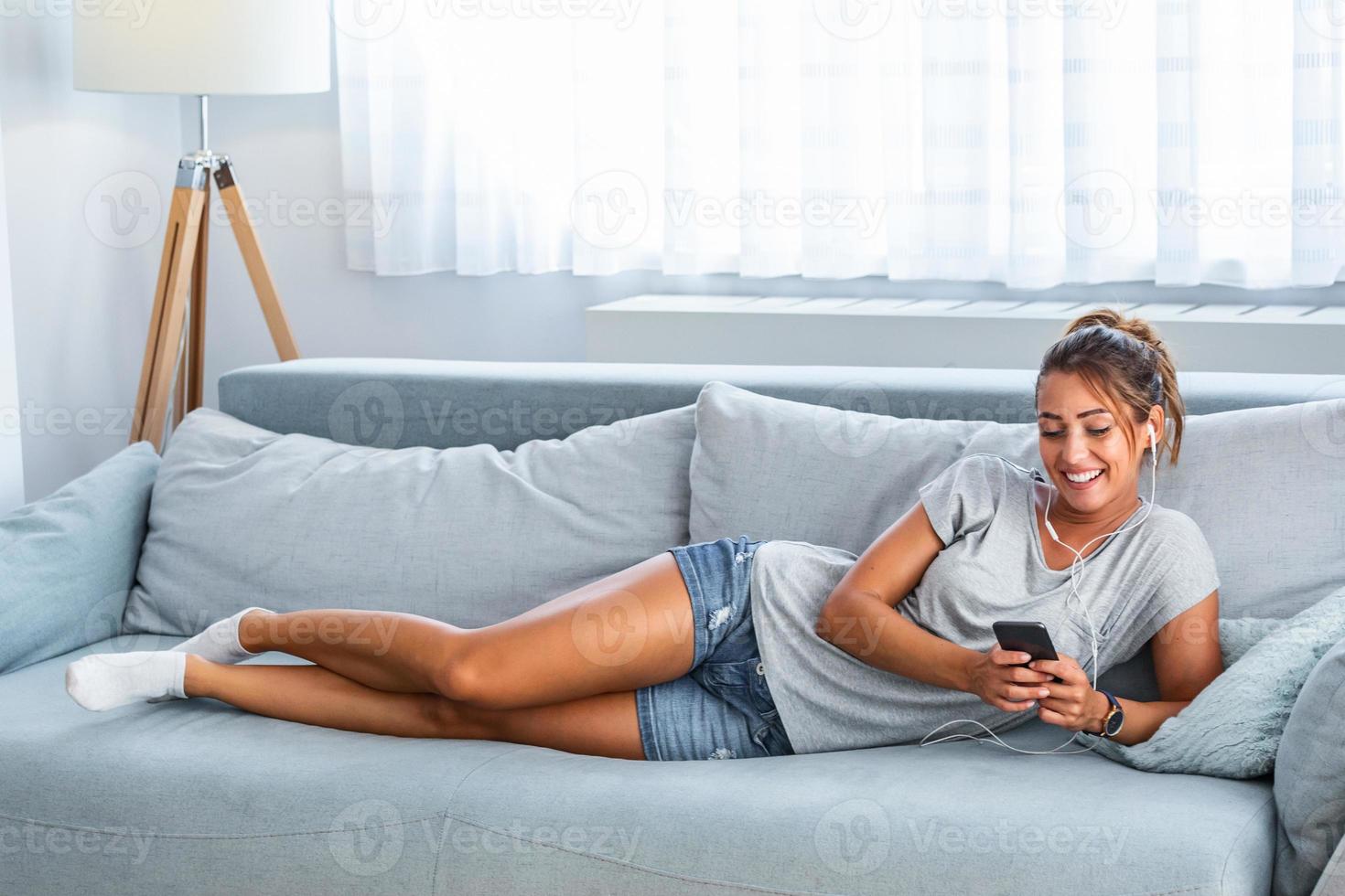 relájate al ritmo de mi música. atractiva mujer joven con auriculares y teléfono inteligente sentado en el sofá en casa. hermosa chica en denim y auriculares está escuchando música usando un teléfono inteligente foto