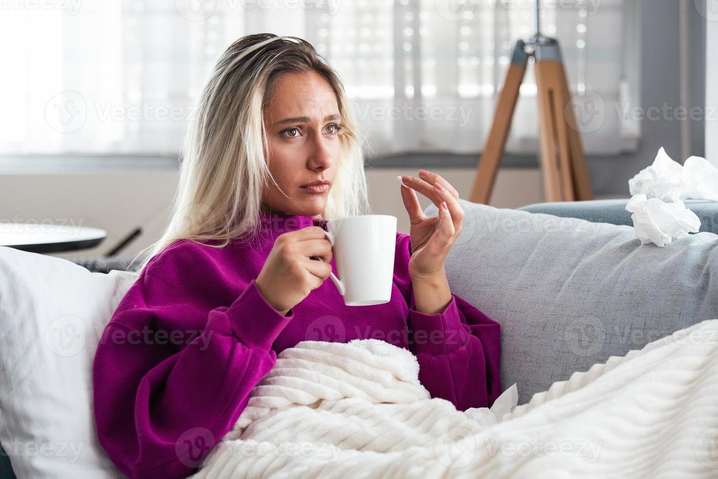 concepto de sufrir un dolor terrible. mujer hermosa, triste, molesta, infeliz, preocupada, débil con cabello rubio, vestida con ropa informal, sentada en un sofá y tomando una pastilla foto