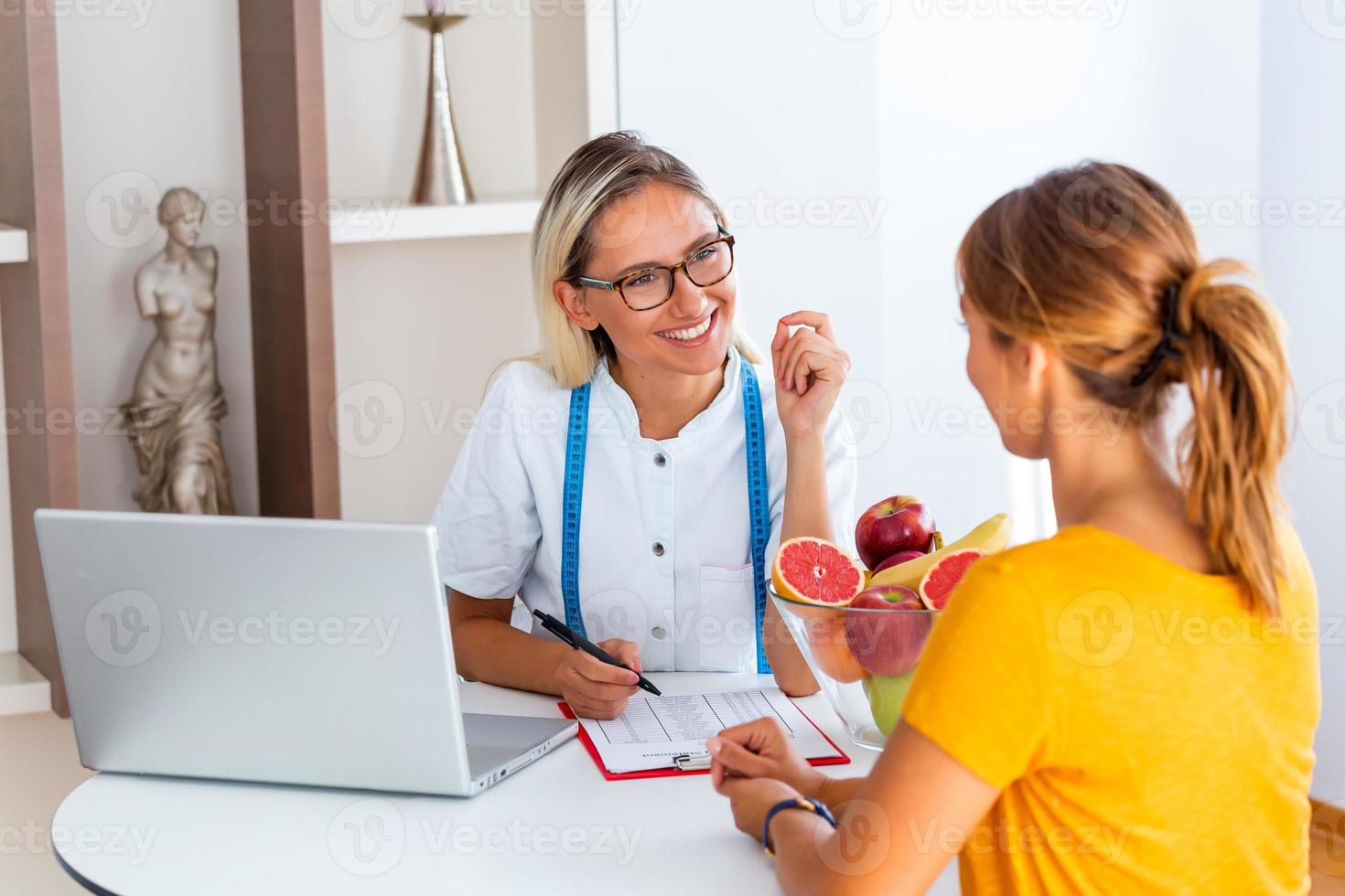 Portrait of young smiling female nutritionist in the consultation room. Making diet plan. Young woman visiting nutritionist in weight loss clinic photo