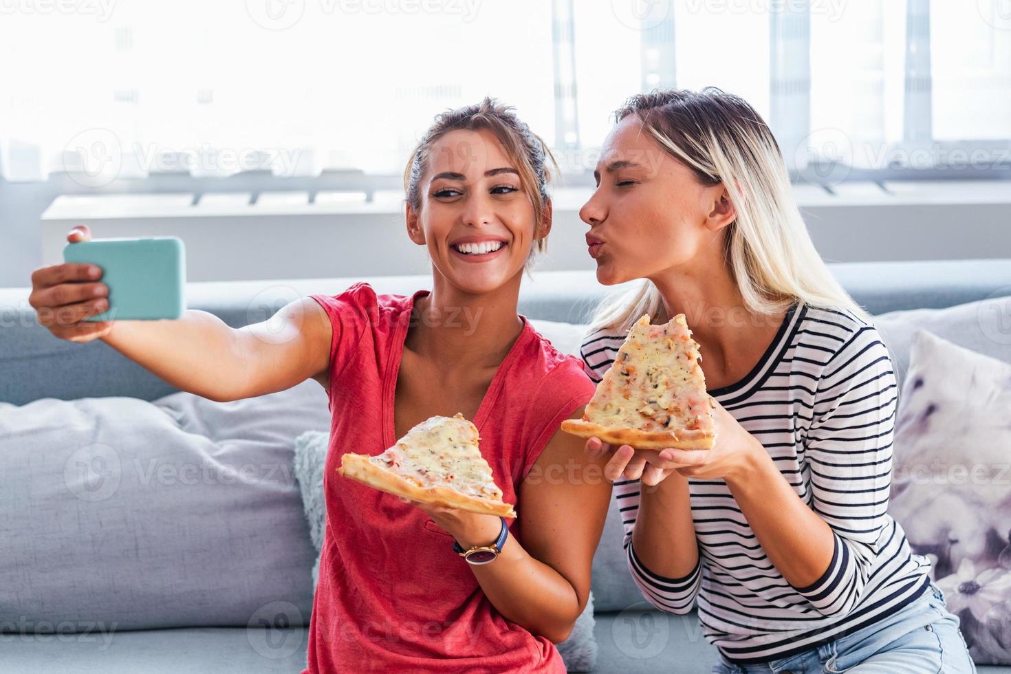 Friends eating pizza and smiling for selfie. They are sharing pizza and making selfie photo on mobile smart phone. They are having party at home, eating pizza and having fun.