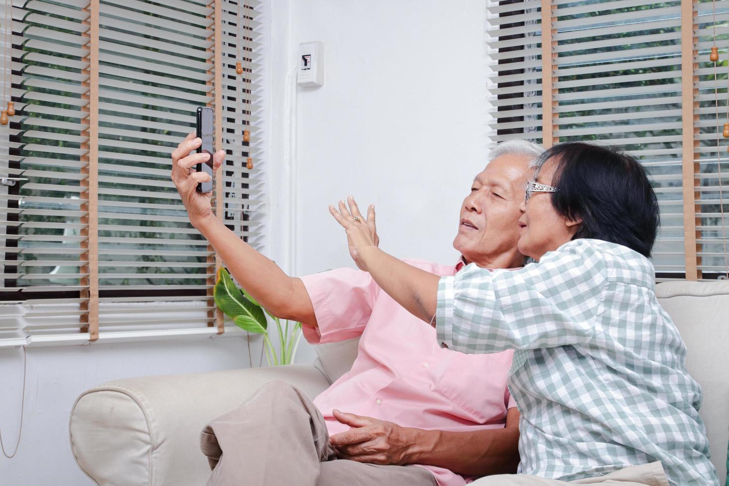 Asian elderly couple Meet in virtual family, children and grandchildren through mobile phones during coronavirus. Social distance. Happy smiling couple sharing their smartphones on the sofa at home. photo