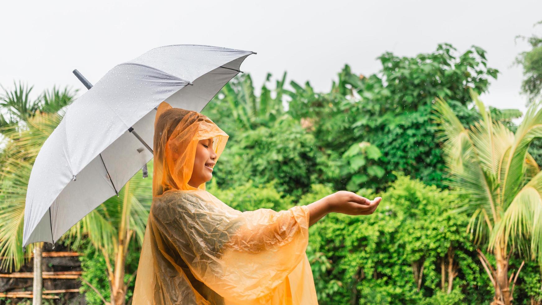 niño asiático con impermeable naranja sosteniendo un paraguas feliz y divirtiéndose bajo la lluvia en un día lluvioso foto