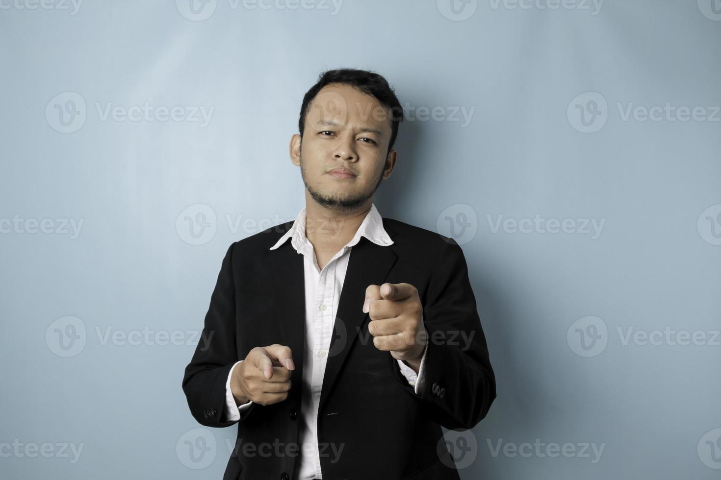 Portrait of young Asian businessman in black suit pointing index finger at camera, posing isolated over blue background. Cheerful smiling guy picking, choosing and indicating photo