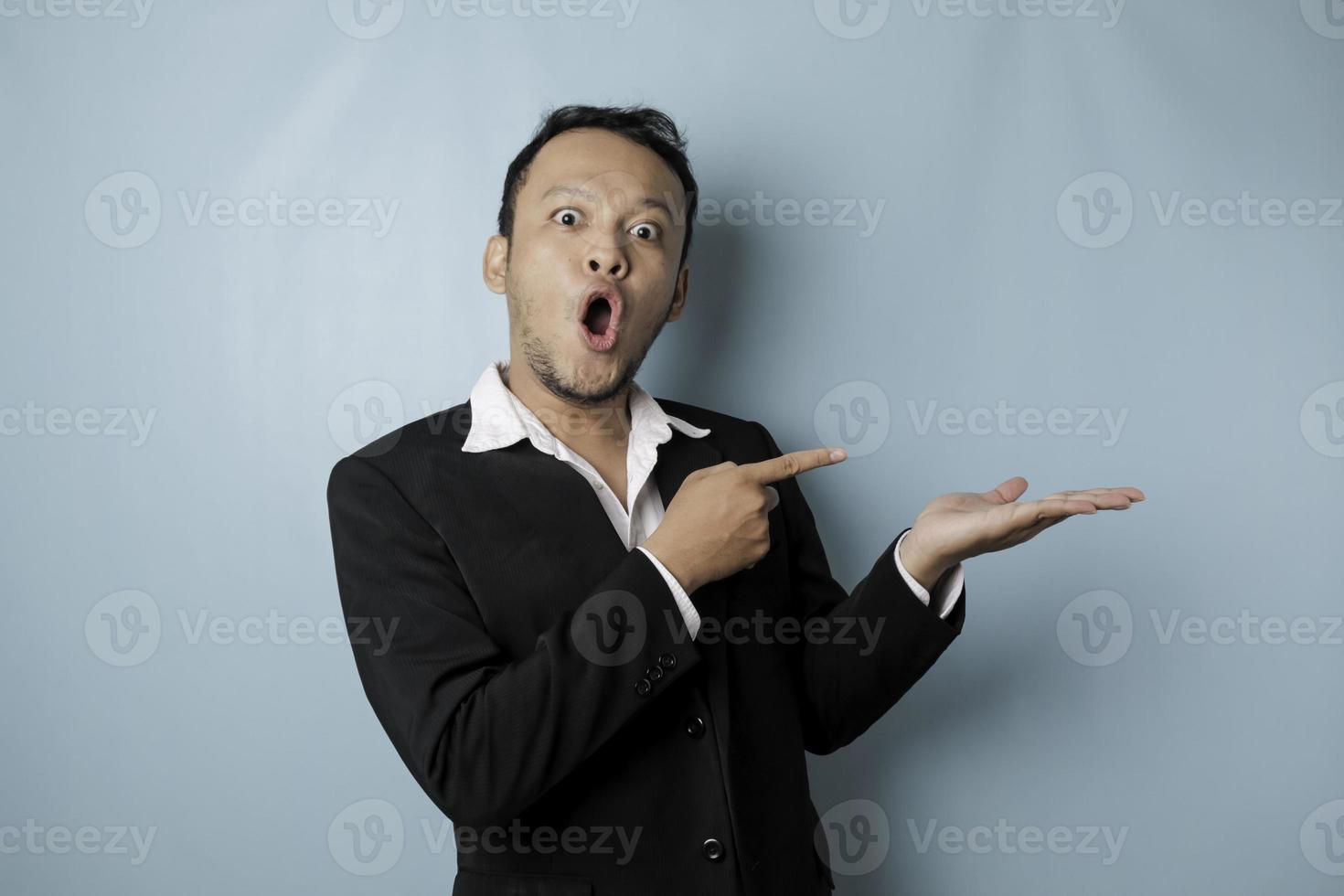 Shocked Asian man wearing suit pointing at the copy space beside him and smiling, isolated by blue background photo