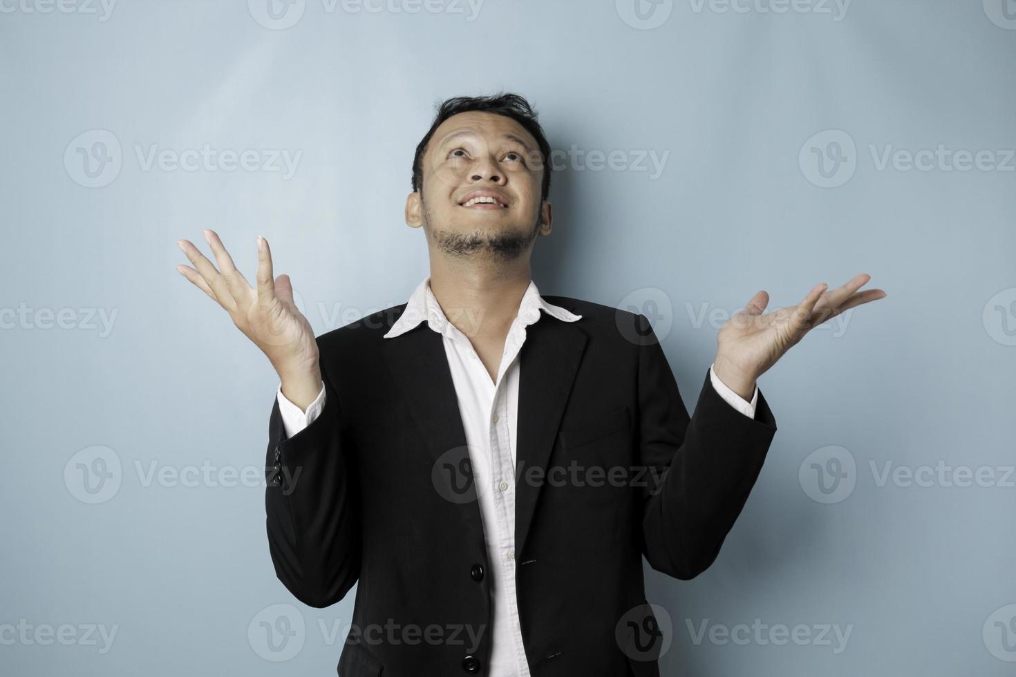 Excited Asian man wearing suit pointing at the copy space upside him, isolated by blue background photo