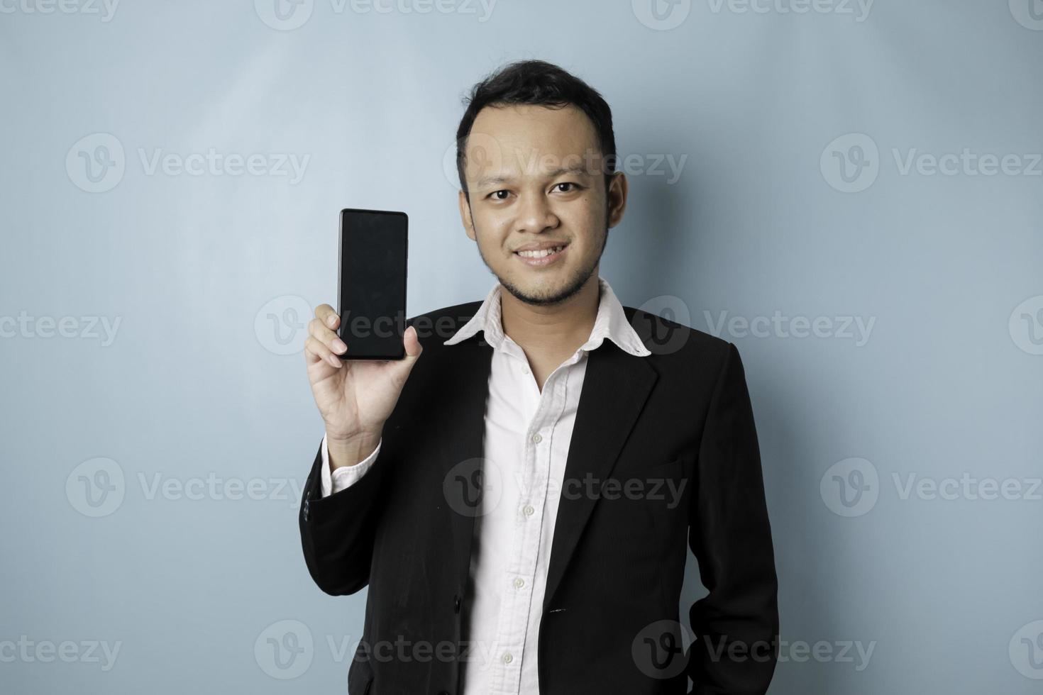 A portrait of a happy Asian businessman is smiling and holding his smartphone showing copy space on it's screen wearing black suit isolated by a blue background photo