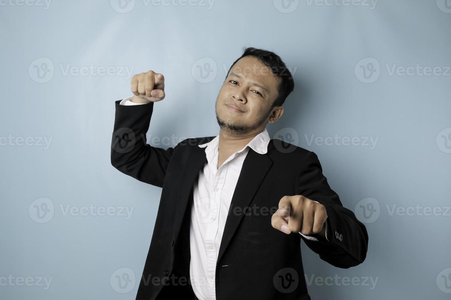 Portrait of young Asian businessman in black suit pointing index finger at camera, posing isolated over blue background. Cheerful smiling guy picking, choosing and indicating photo