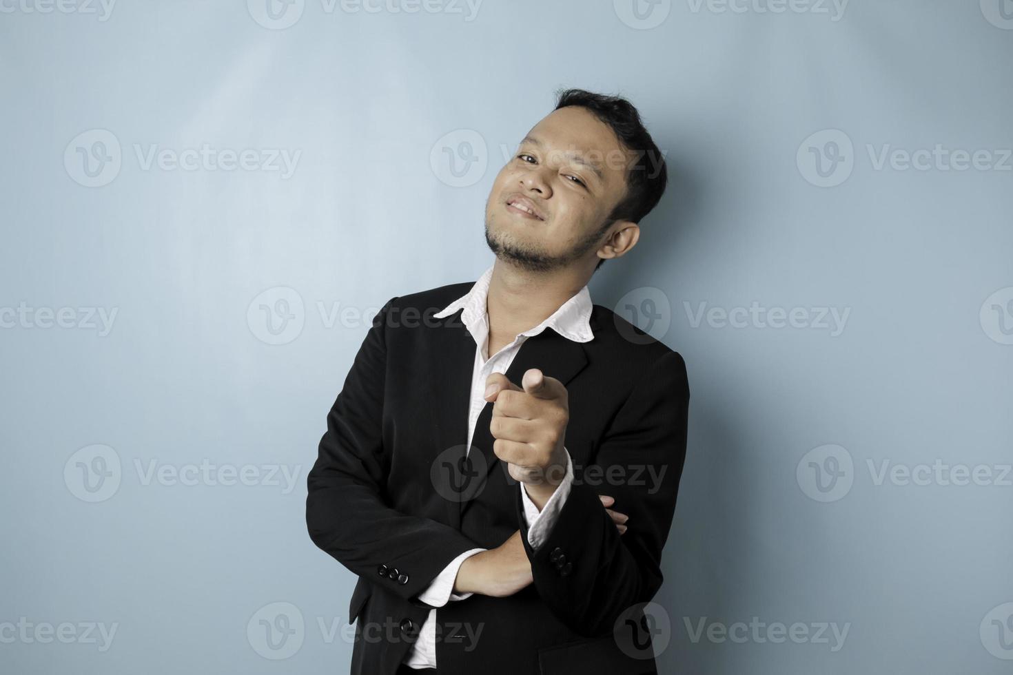 Portrait of young Asian businessman in black suit pointing index finger at camera, posing isolated over blue background. Cheerful smiling guy picking, choosing and indicating photo