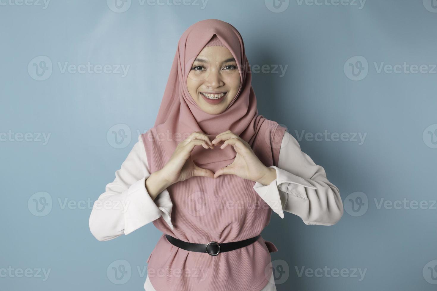 An attractive young Asian Muslim woman wearing a pink hijab feels happy and a romantic shapes heart gesture expresses tender feelings photo