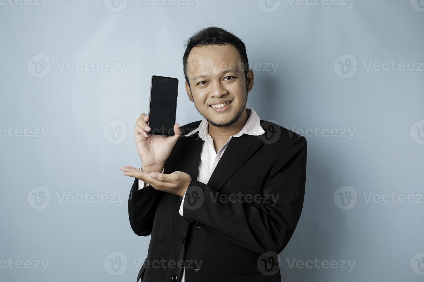 un retrato de un hombre de negocios asiático feliz sonríe y sostiene su teléfono inteligente que muestra espacio de copia en su pantalla con traje negro aislado por un fondo azul foto