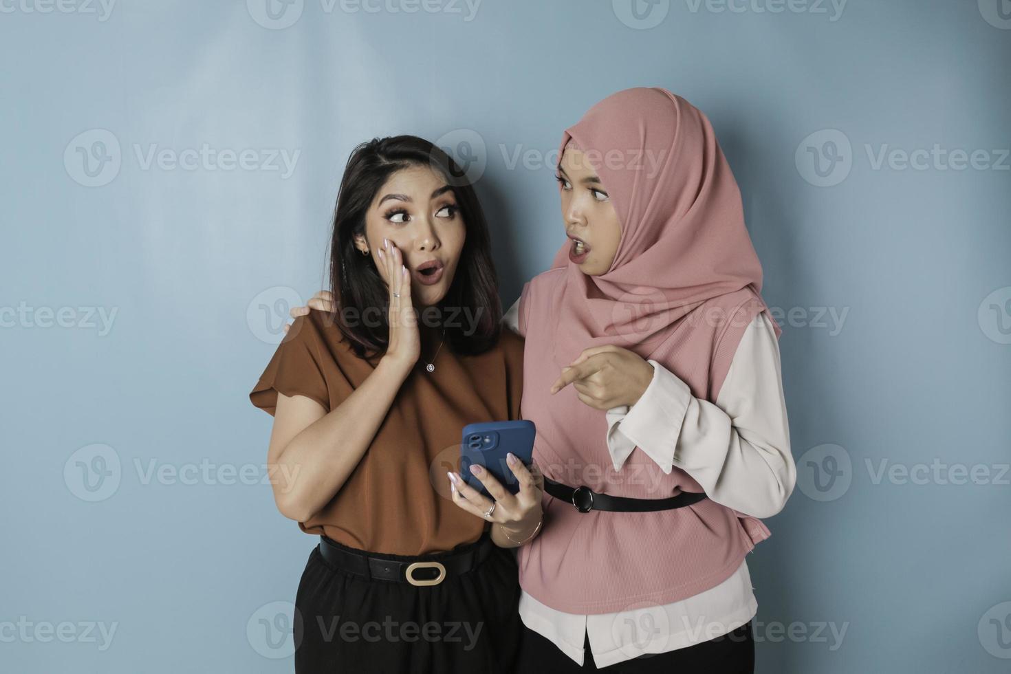 Two surprised Asian women holding smartphone, isolated by blue background photo