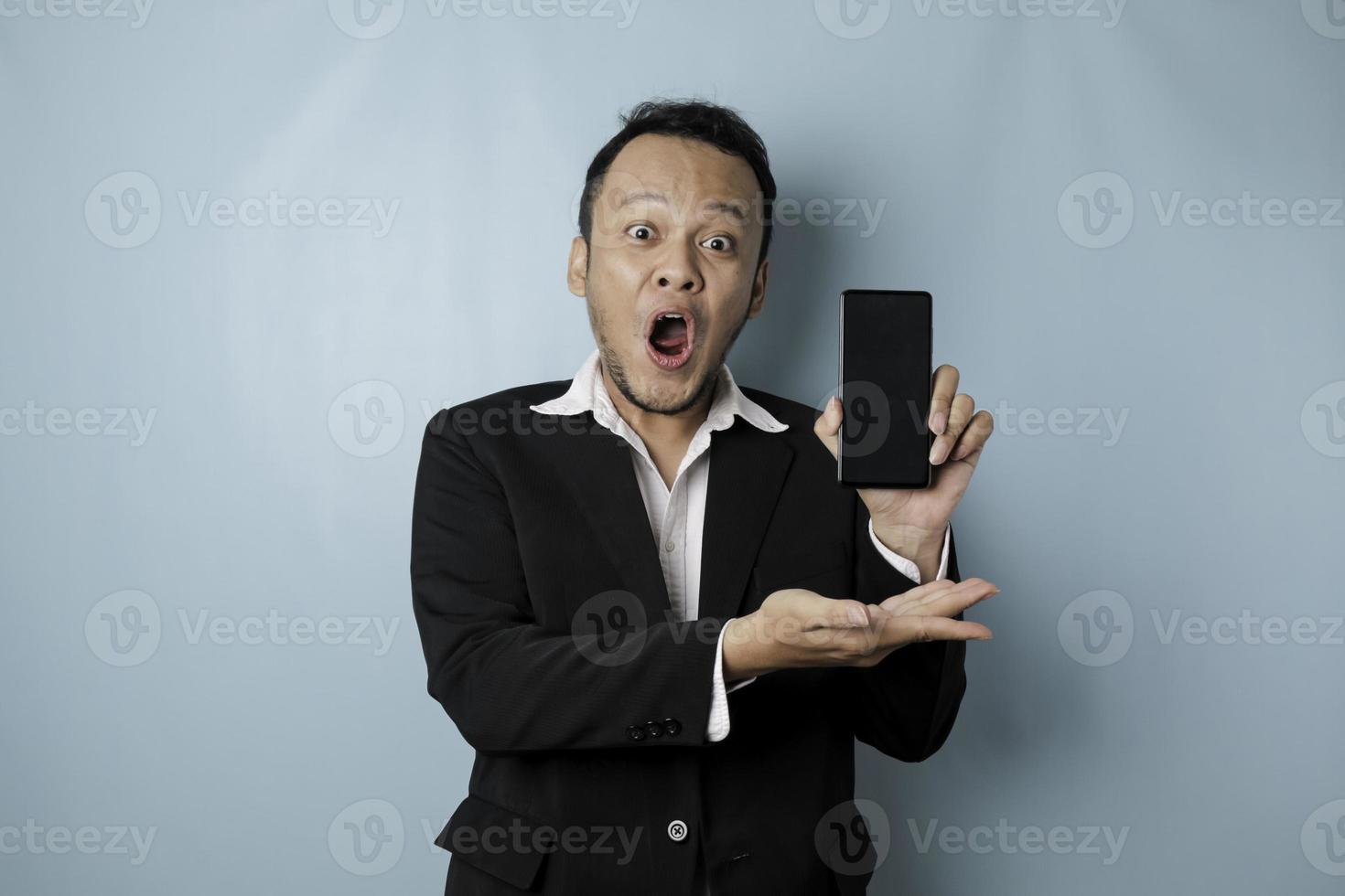 Surprised Asian businessman wearing a black suit holding his smartphone and showing copy space on it's screen, isolated by a blue background photo