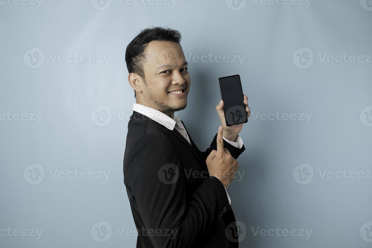 A portrait of a happy Asian businessman is smiling and holding his smartphone showing copy space on it's screen wearing black suit isolated by a blue background photo