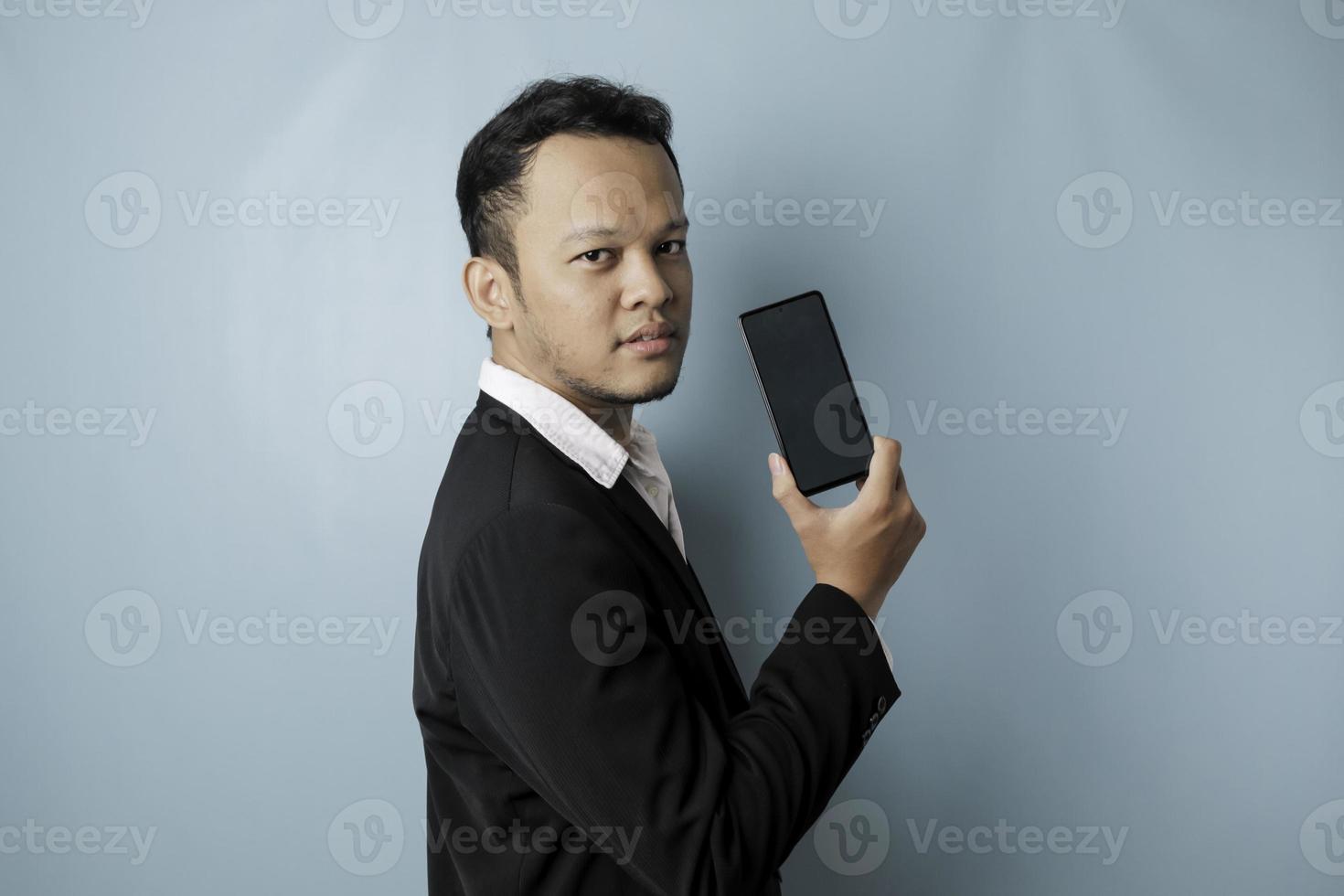 un joven hombre de negocios asiático con traje negro se siente serio y se enfoca sosteniendo un teléfono inteligente y mostrando el espacio de copia en su pantalla en la mano. foto