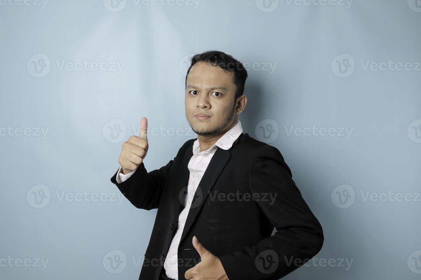 Excited Asian businessman wearing suit gives thumbs up hand gesture of approval, isolated by blue background photo