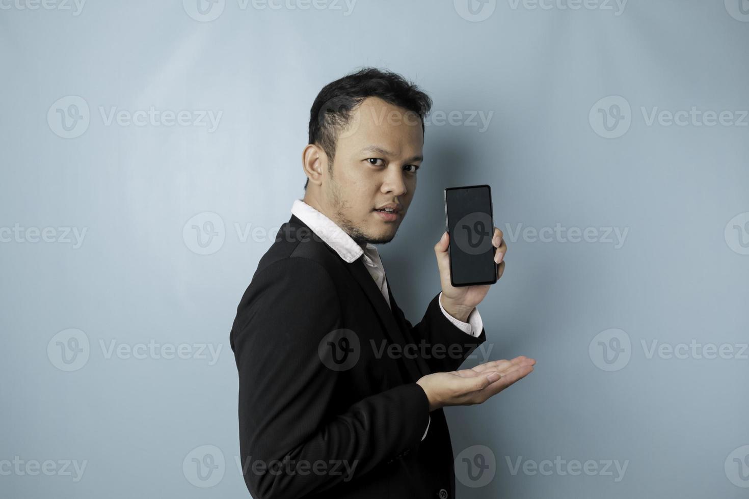 un retrato de un hombre de negocios asiático feliz sonríe y sostiene su teléfono inteligente que muestra espacio de copia en su pantalla con traje negro aislado por un fondo azul foto