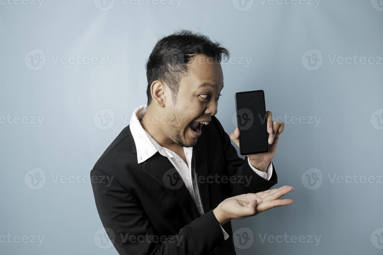 Surprised Asian businessman wearing a black suit holding his smartphone and showing copy space on it's screen, isolated by a blue background photo