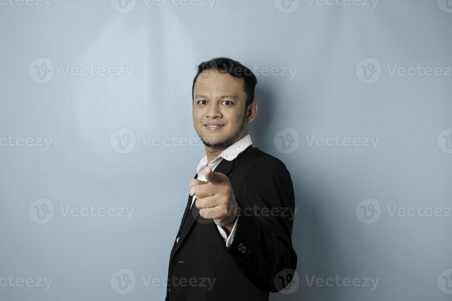 Portrait of young Asian businessman in black suit pointing index finger at camera, posing isolated over blue background. Cheerful smiling guy picking, choosing and indicating photo