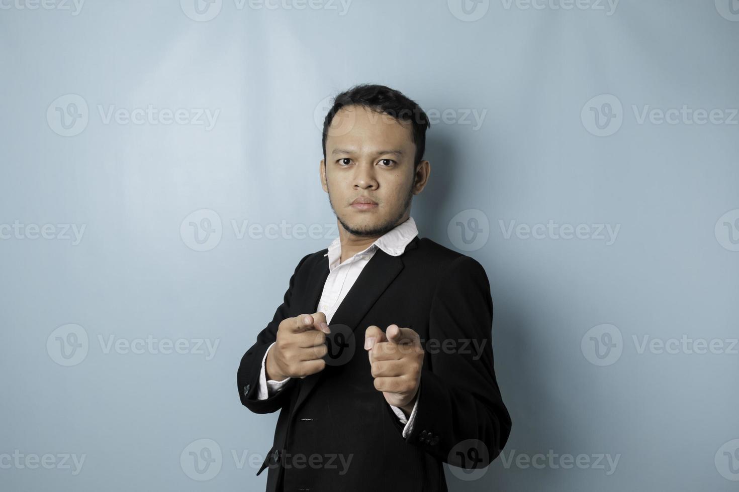 Portrait of young Asian businessman in black suit pointing index finger at camera, posing isolated over blue background. Cheerful smiling guy picking, choosing and indicating photo