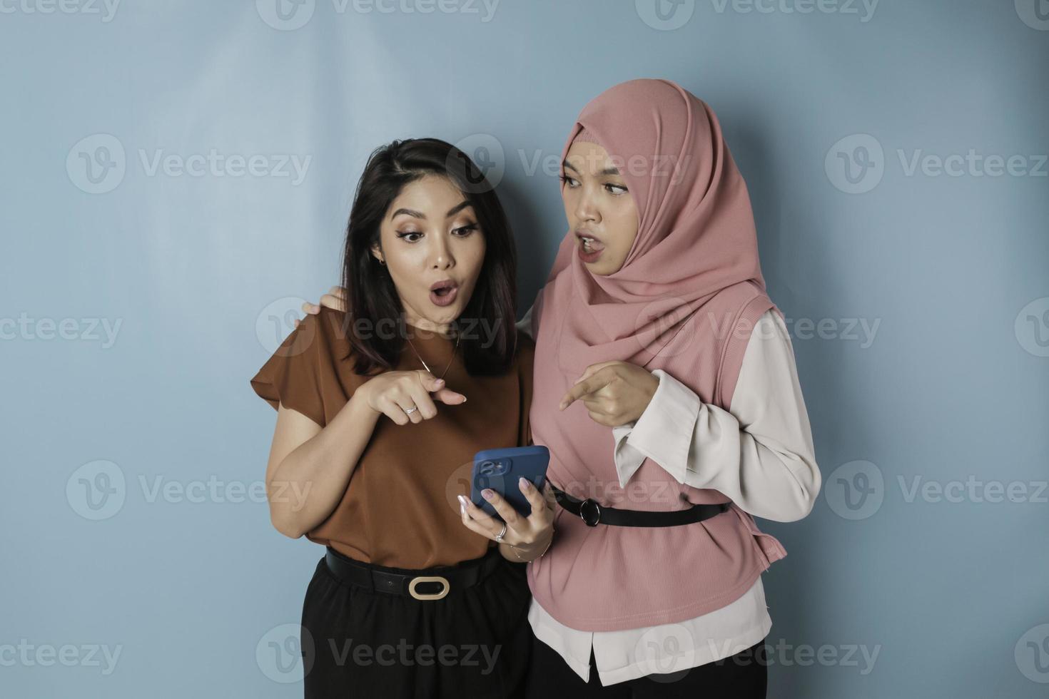 Two surprised Asian women holding smartphone, isolated by blue background photo