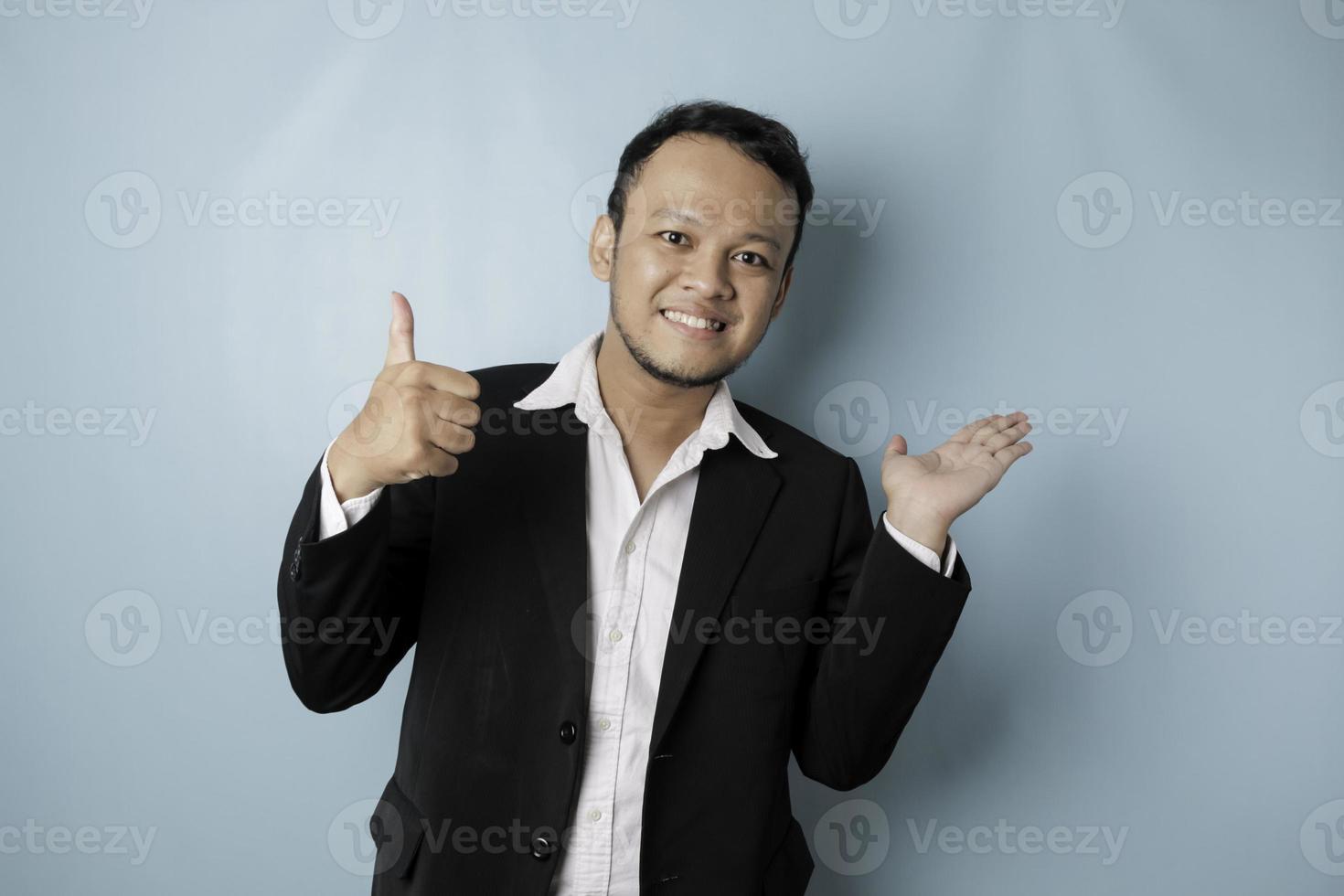 Excited Asian man wearing suit pointing at the copy space beside him and smiling, isolated by blue background photo