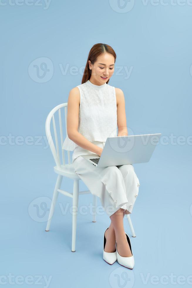 Fashionable young girl sitting on a chair with laptop isolated on blue background photo