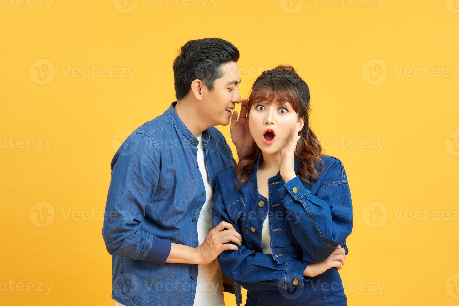Portrait of asian man whispering secret or interesting gossip to excited woman in her ear isolated over yellow background photo